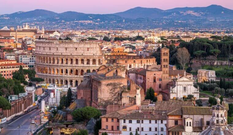 Skyline of Rome, Italy at sunset, with historic buildings including the Colosseum in the foreground, and mountains in the background