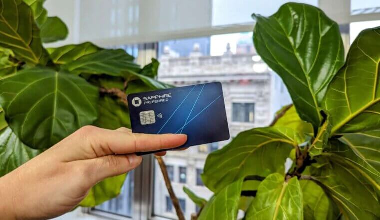 The Chase Sapphire Preferred Card held up before a leafy plant with buildings seen through the window in the background
